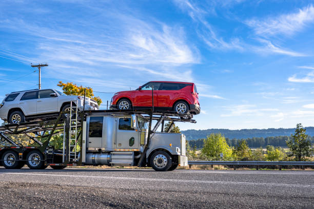 Industrial grade classic big rig car hauler white semi truck tractor transporting cars on the modular hydraulic semi trailer driving on the narrow highway road with trees on the side