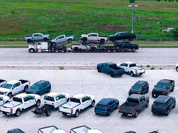Drone mid-low photograph showing hundreds of cars and trucks organized in neat rows at automotive dealership or vehicle storage facility. The vast parking lot demonstrates the scale of modern automotive retail operations with various vehicle types including pickup trucks, SUVs, and sedans in multiple colors arranged systematically across the concrete pavement.